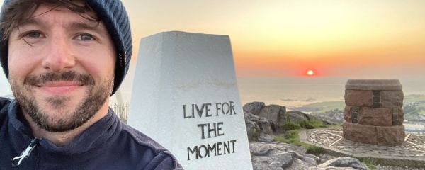 Chris is standing outdoors on a rocky hilltop at sunset. In the foreground, there is a white stone monument with the engraved words “LIVE FOR THE MOMENT.” A circular stone structure is visible to the right, surrounded by a decorative patterned platform. The landscape stretches out toward the coastline below, with a glowing orange sun setting over the distant water and casting warm light across the sky.